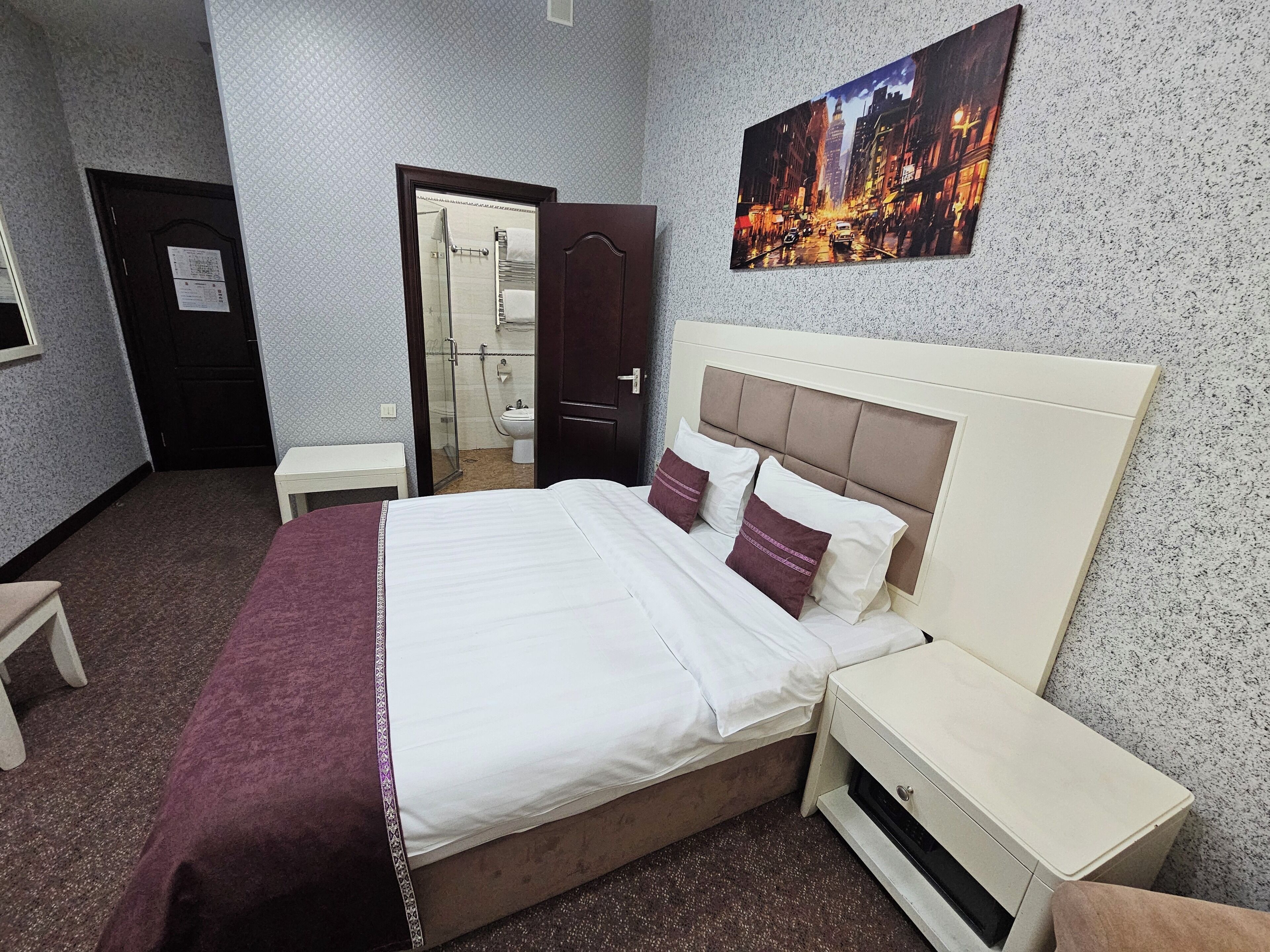 A hotel room with a bed, a nightstand, a small table, a bathroom visible through an open door, and a cityscape painting on the wall.