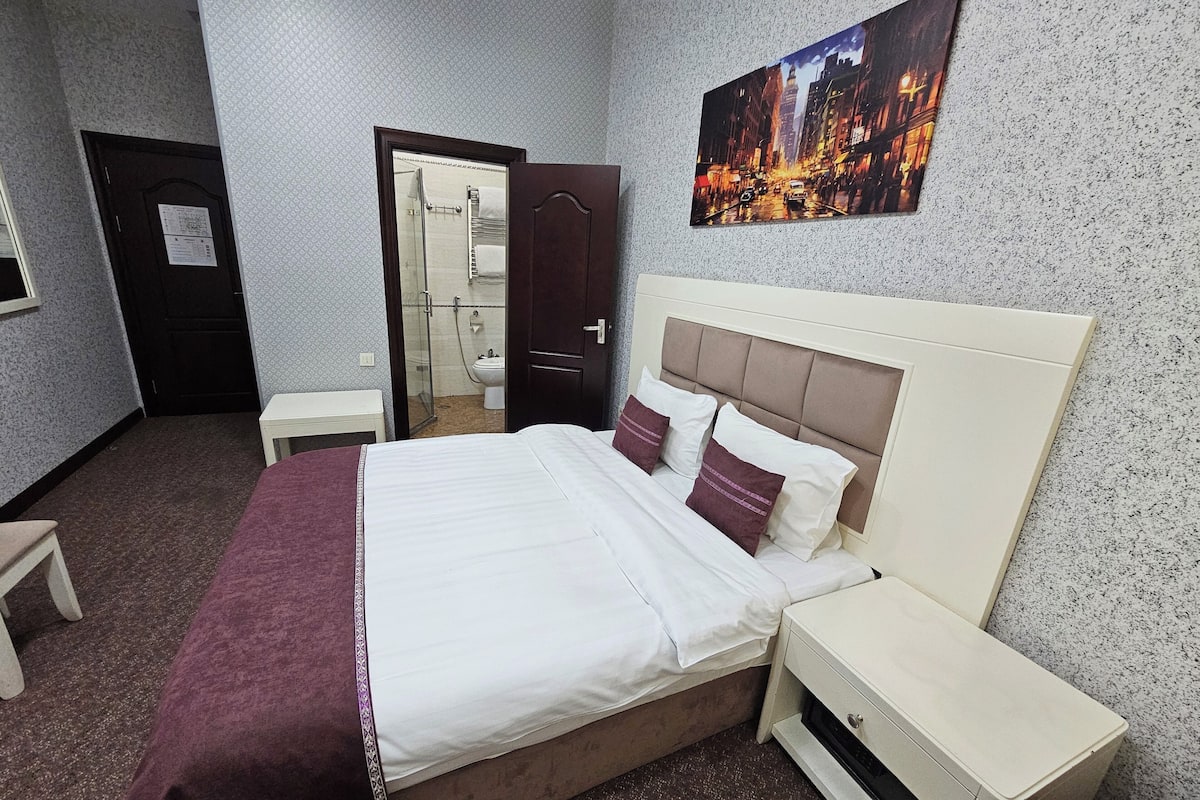 A hotel room with a bed, a nightstand, a small table, a bathroom visible through an open door, and a cityscape painting on the wall.