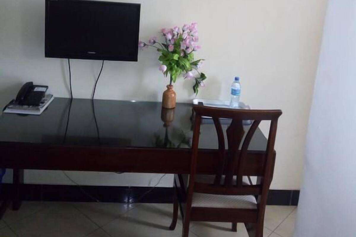 A room with a flat-screen TV mounted on the wall, a wooden table with a vase of flowers, a chair, and a water bottle on the table.