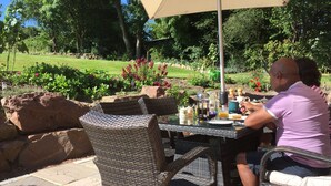 Un patio con sillas de mimbre y una mesa, rodeado de un jardín con rocas y plantas.