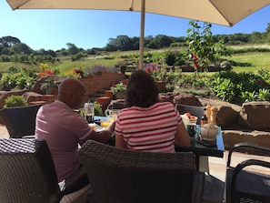 A couple dining at an outdoor table with a view of a landscaped garden and hills. - Martlets B&B (Shanklin)