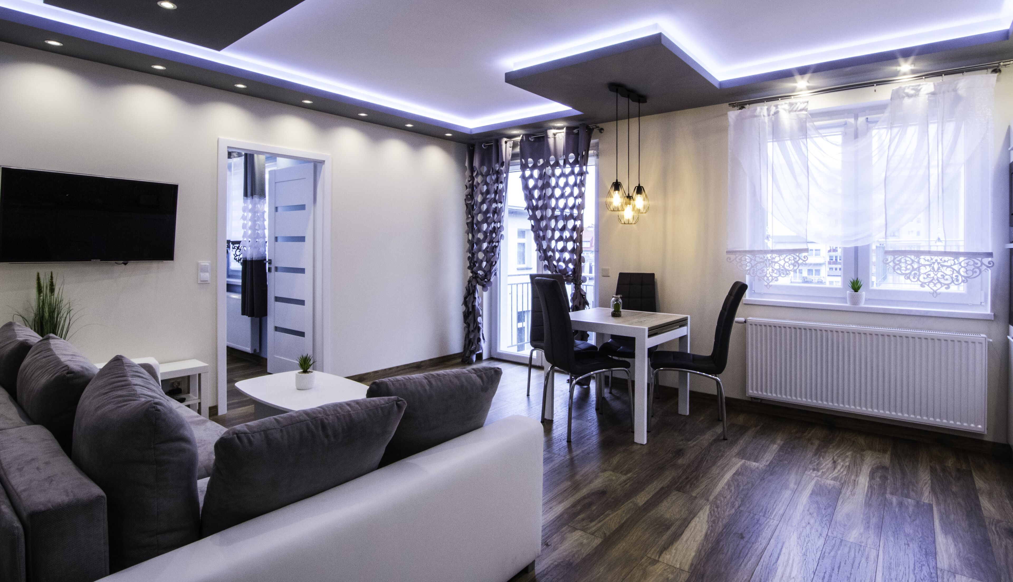 A modern living room with a sofa, a dining table with chairs, a TV mounted on the wall, and a window with curtains.