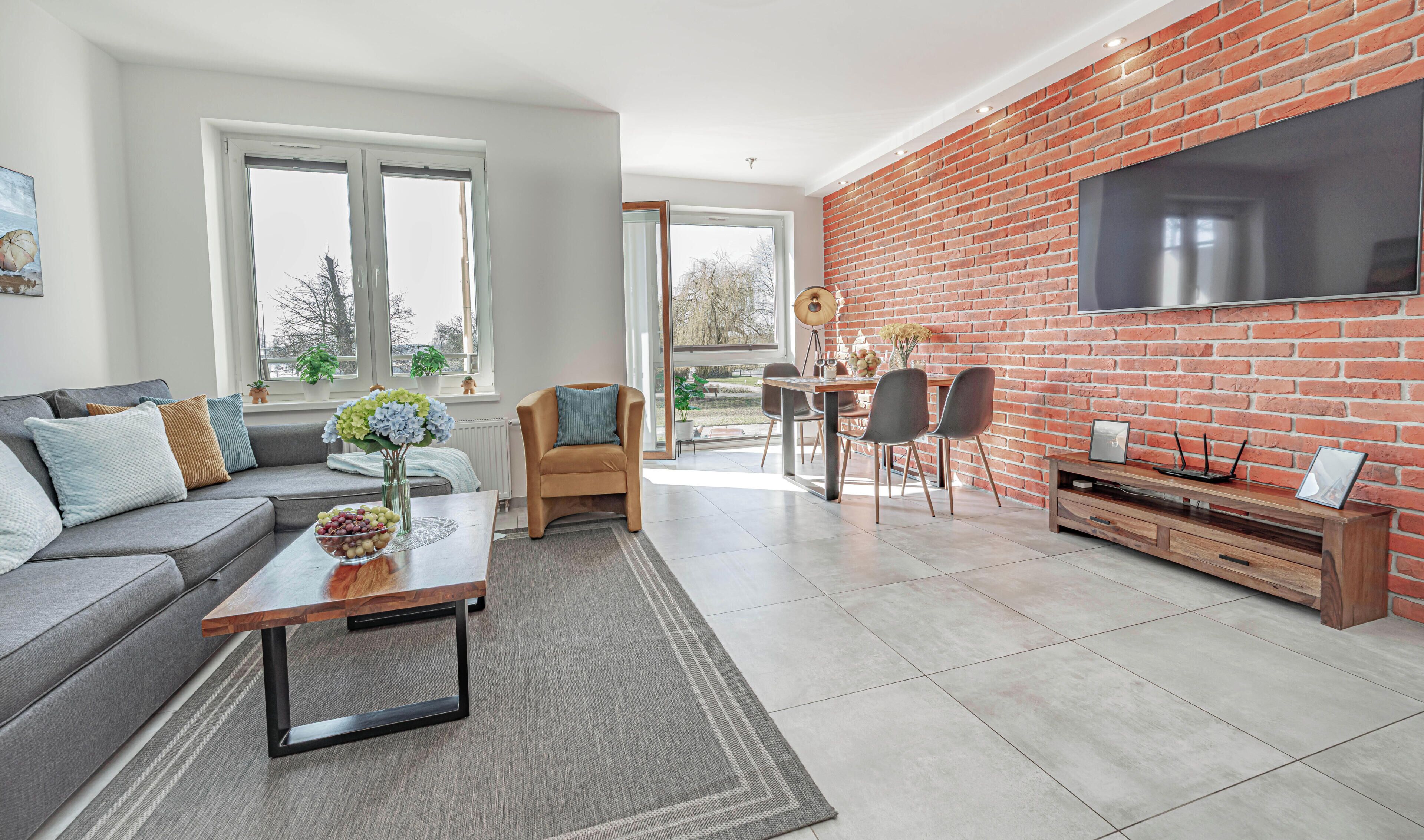 A modern living room with a grey sofa, a coffee table, a brick accent wall, and a large TV.