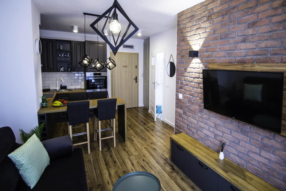 Una cocina moderna con armarios oscuros, un comedor con mesa y sillas de madera, y una sala de estar con sofá negro y televisor montado en una pared de ladrillo.