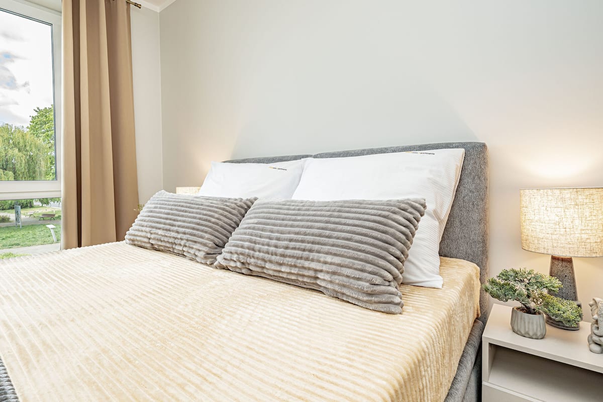 Una cama bien tendida con un edredón color beige y dos almohadas, una lámpara de noche, una pequeña planta en maceta y una figurita decorativa de un elefante sobre una mesita de noche.