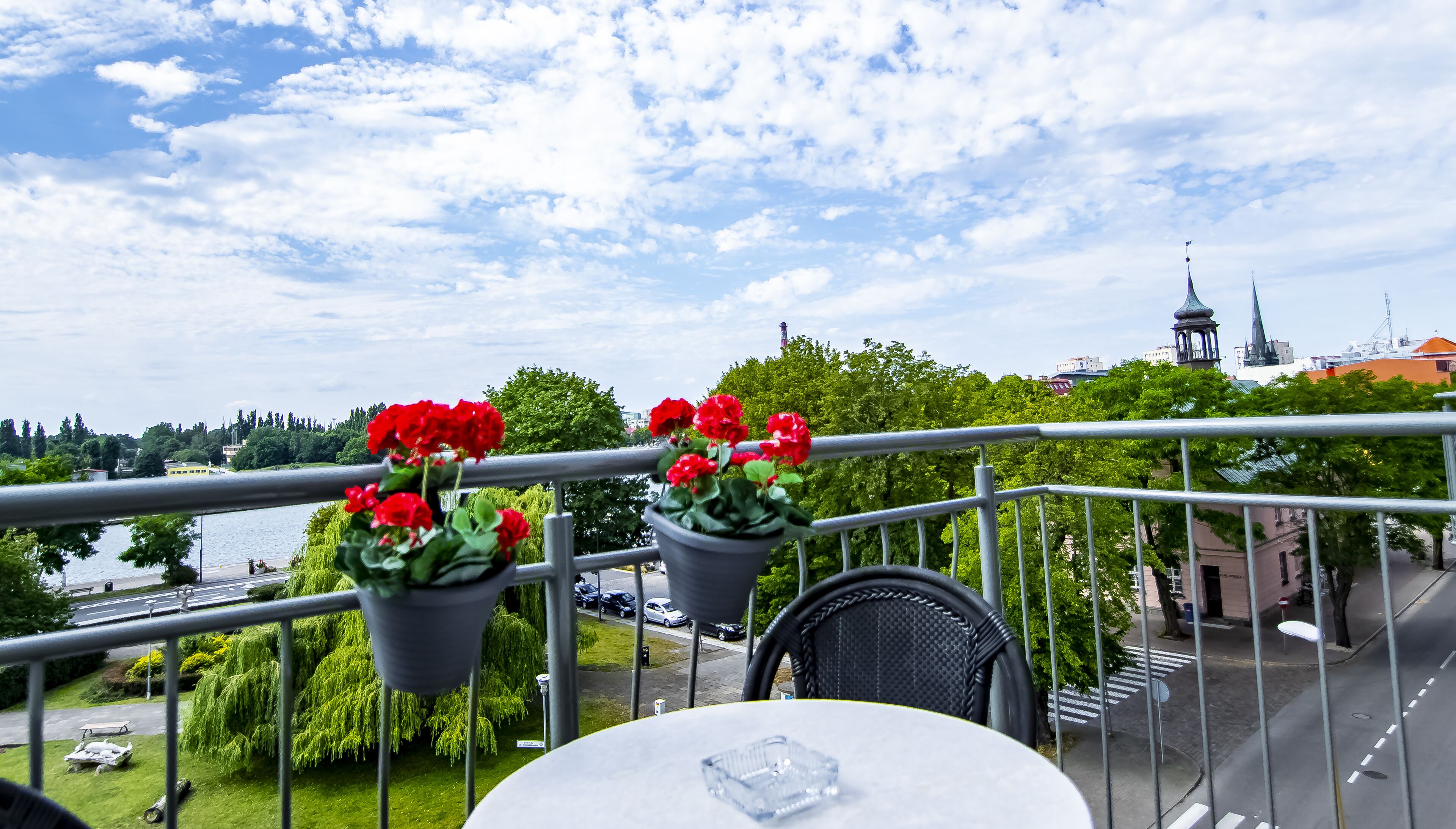 A balcony with potted red flowers, a view of a river, and a cityscape in the background.