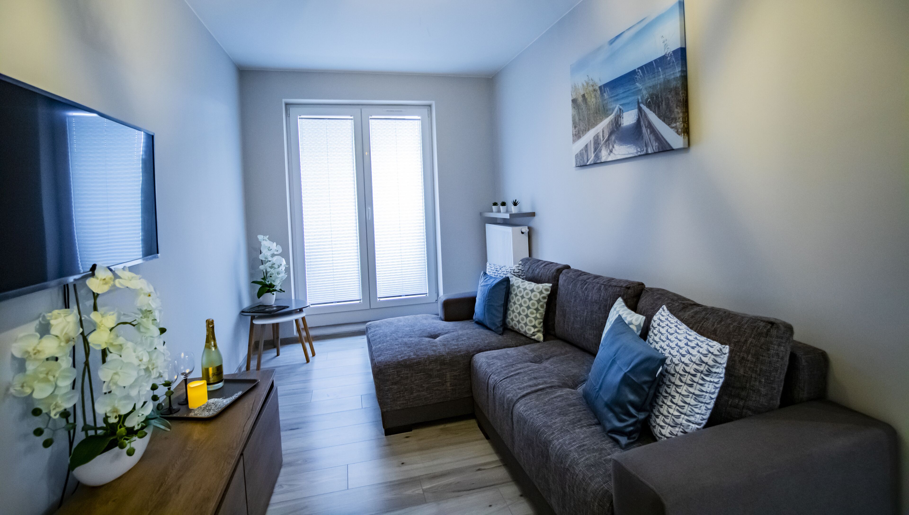 A modern living room with a grey sofa, a small side table with a vase of white flowers, and a painting on the wall.