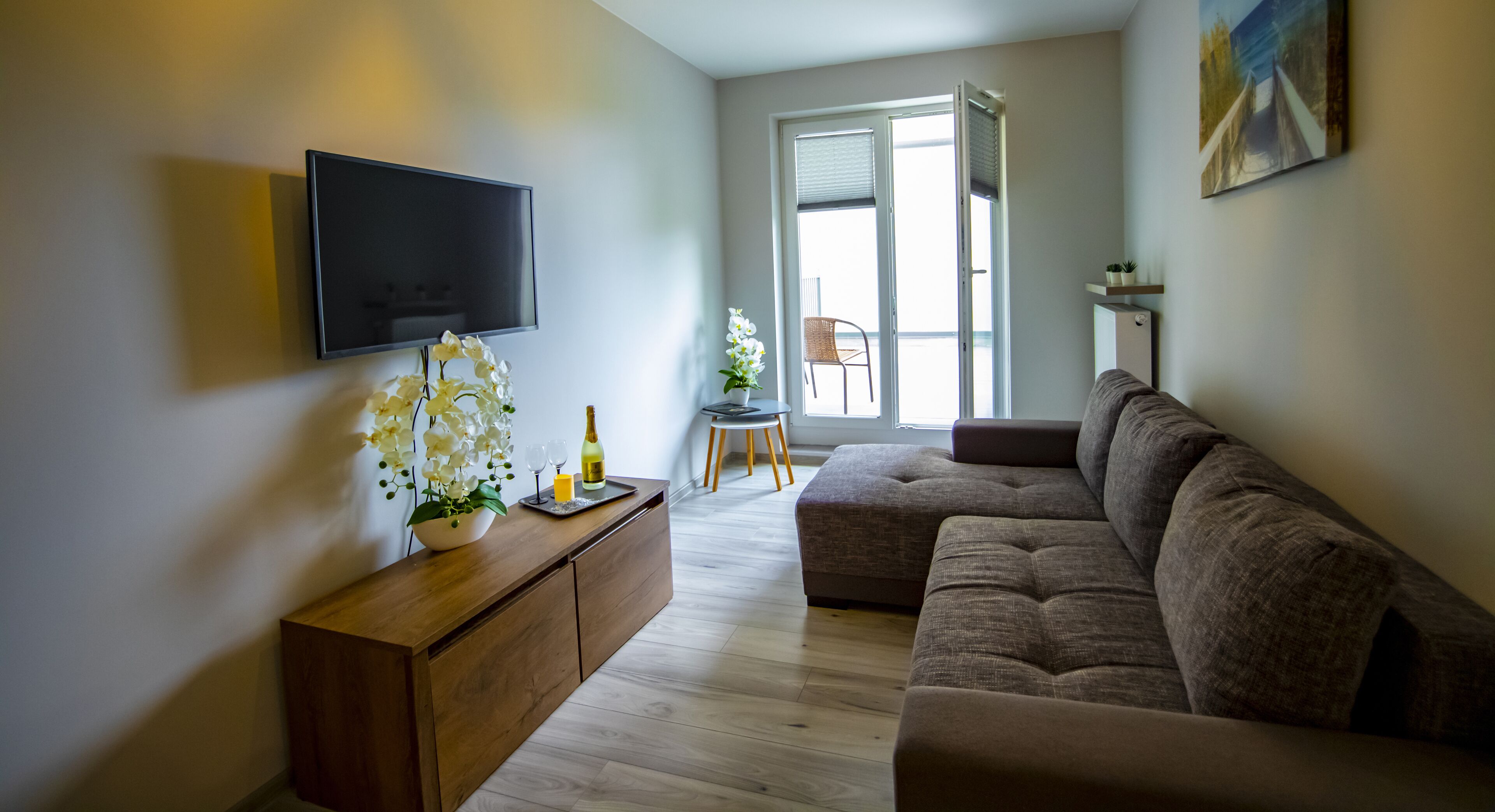 A modern living room with a flat-screen TV, a wooden console table with a vase of flowers, a brown sectional sofa, and a dining area with a chair and table.
