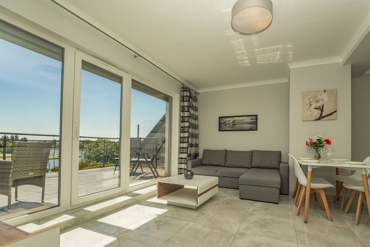 Una sala moderna con un sofá gris, una mesa de comedor con sillas y un ventanal que ofrece vistas a un balcón y zona exterior.