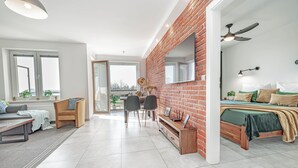 A modern living room with a dining area, a bed with a green bedspread, and a brick accent wall. - Baltic Apartments - Apartamenty Bałtyk (Swinoujscie)