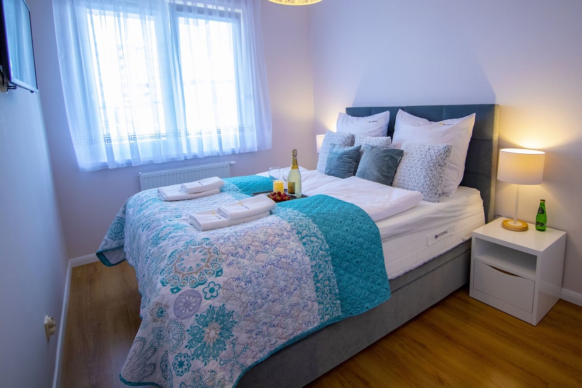Una cama bien hecha con una colcha estampada, una mesita de noche con una botella y un vaso, y una ventana con cortinas traslúcidas.