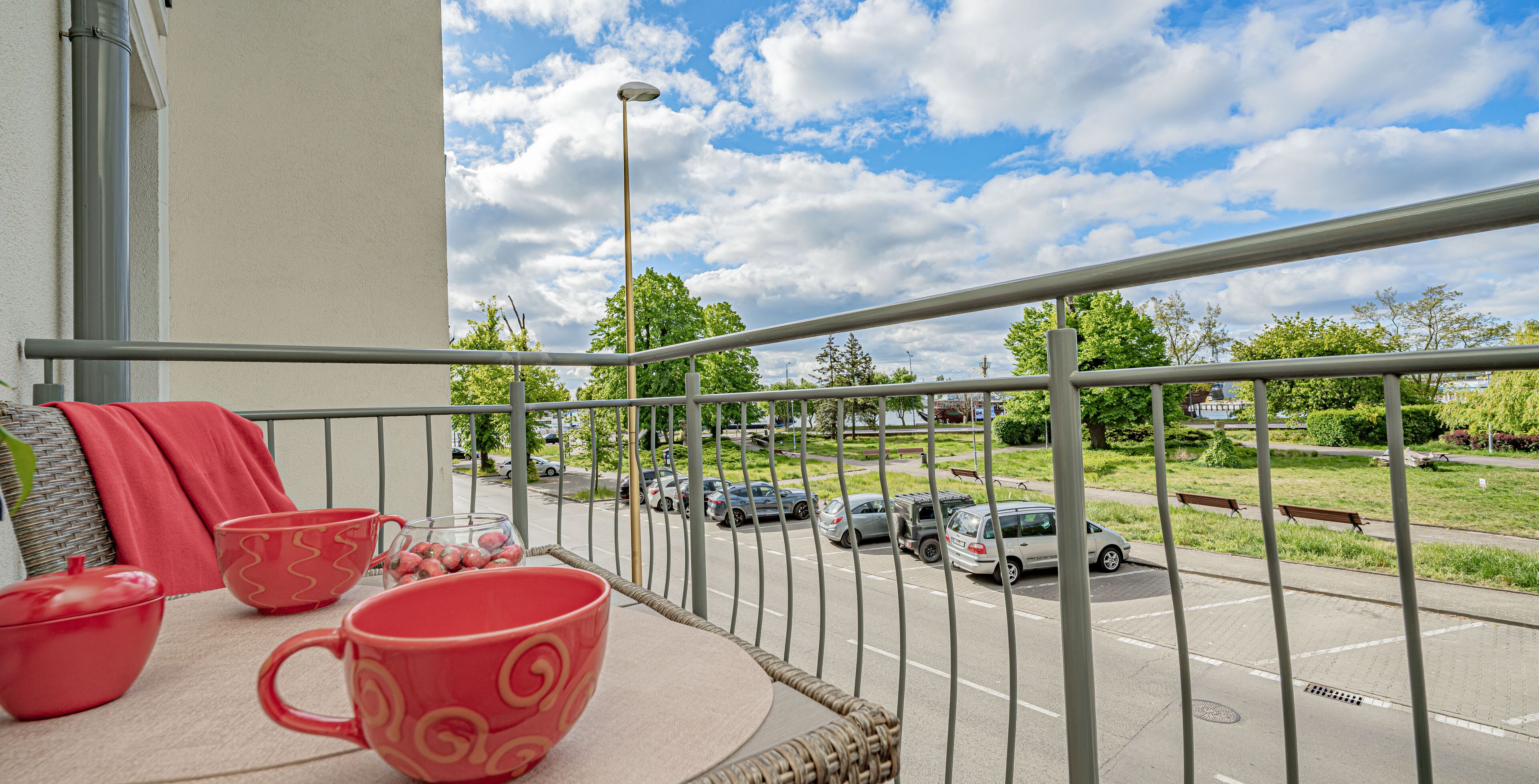 Un balcon agrémenté de meubles en osier, d’un coussin rouge et offrant une vue sur un parking et une rue.