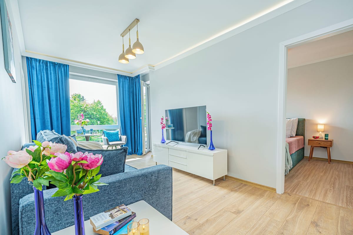 Una sala de estar moderna con sofá, una mesa de centro con libros y flores, televisor y vistas al exterior.