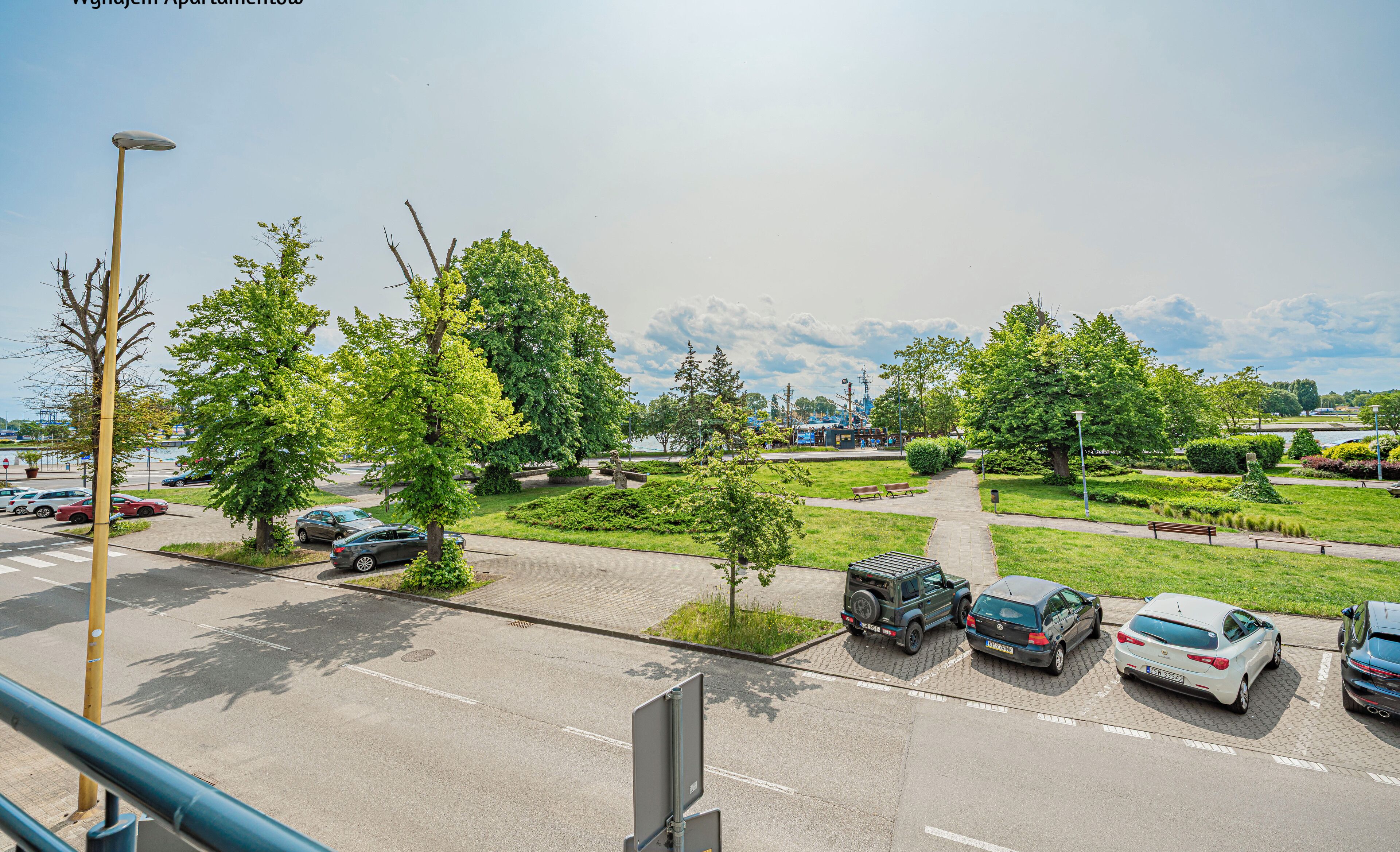 Un parking avec plusieurs voitures, un chemin et des espaces verts avec des arbres.