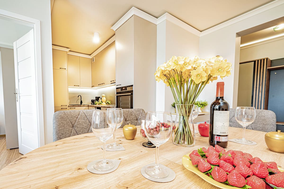 Una mesa de comedor dispuesta con copas de vino, una botella de vino y un plato de fresas en una cocina moderna con sofá y zona de comedor.