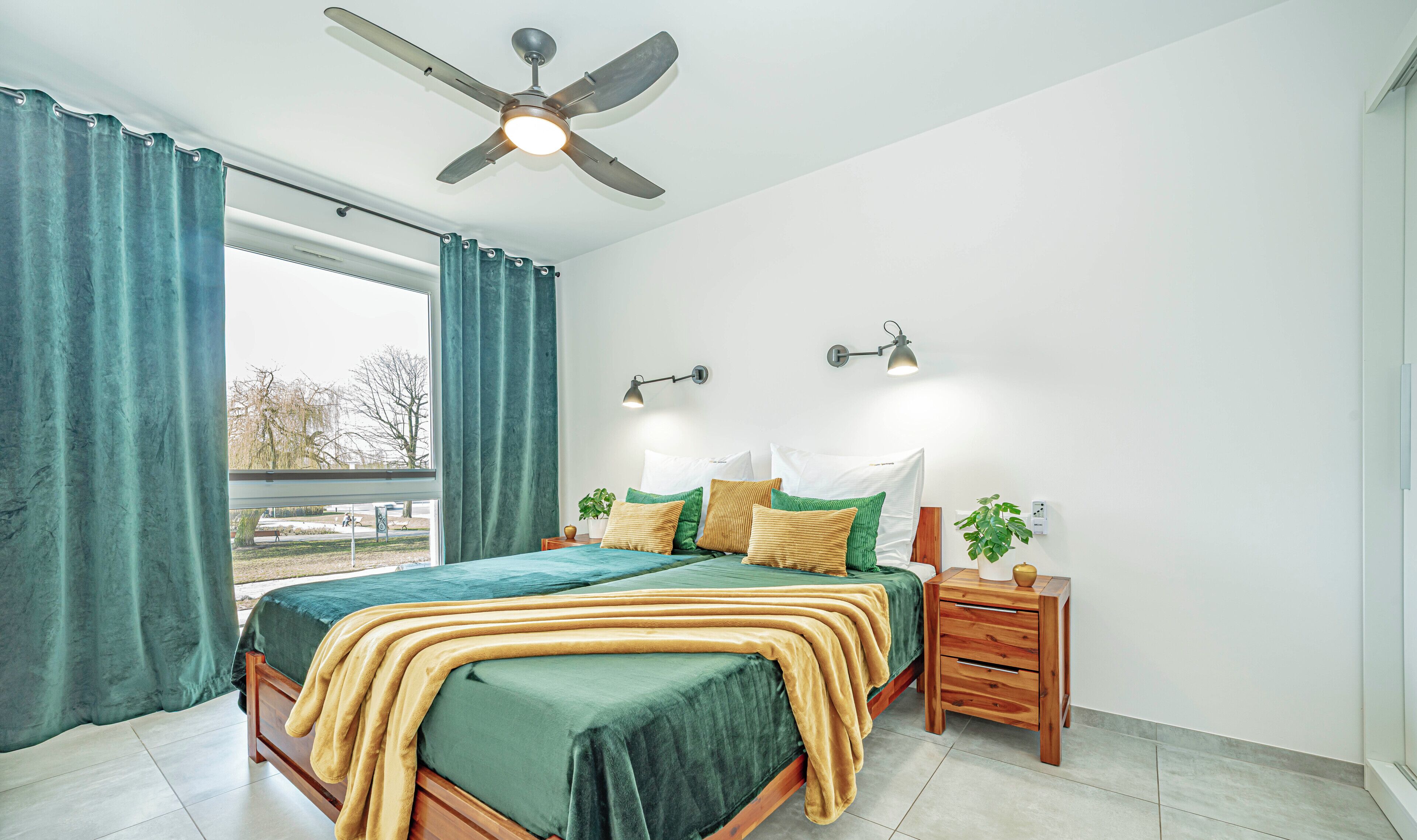 A bedroom with a bed, a nightstand, a ceiling fan, two wall-mounted lamps, and a window with curtains.