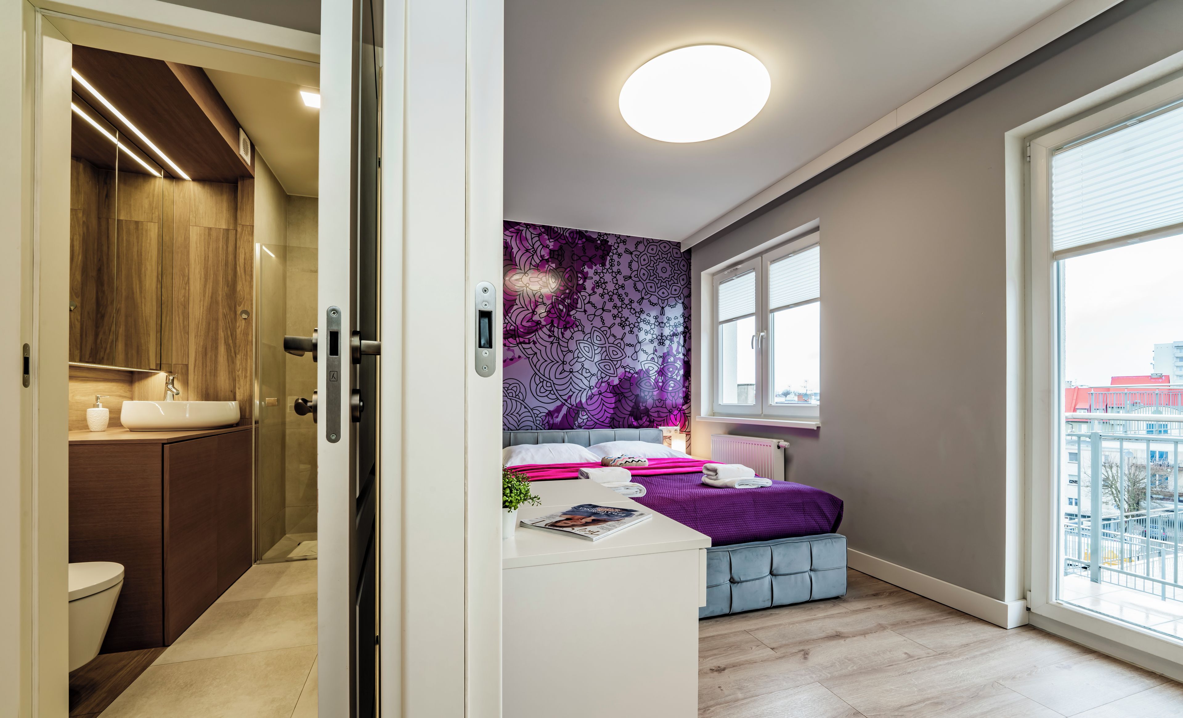 A modern bedroom with a bed, a bedside table, a window, and a bathroom with a toilet and sink.
