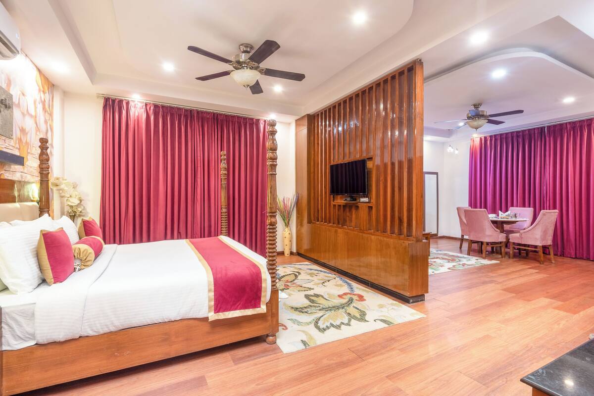 A hotel room with a wooden bed, red curtains, a ceiling fan, a flat-screen TV, and a small dining area with pink chairs.