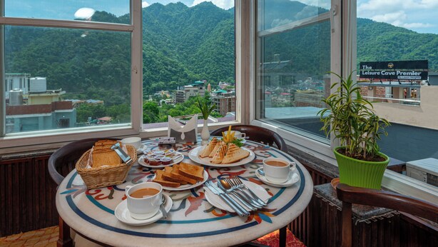 A dining table with plates of food, cups, and utensils, set against a backdrop of a mountainous landscape through large windows. - DLS Hotels on the Ganges (Narendranagar)
