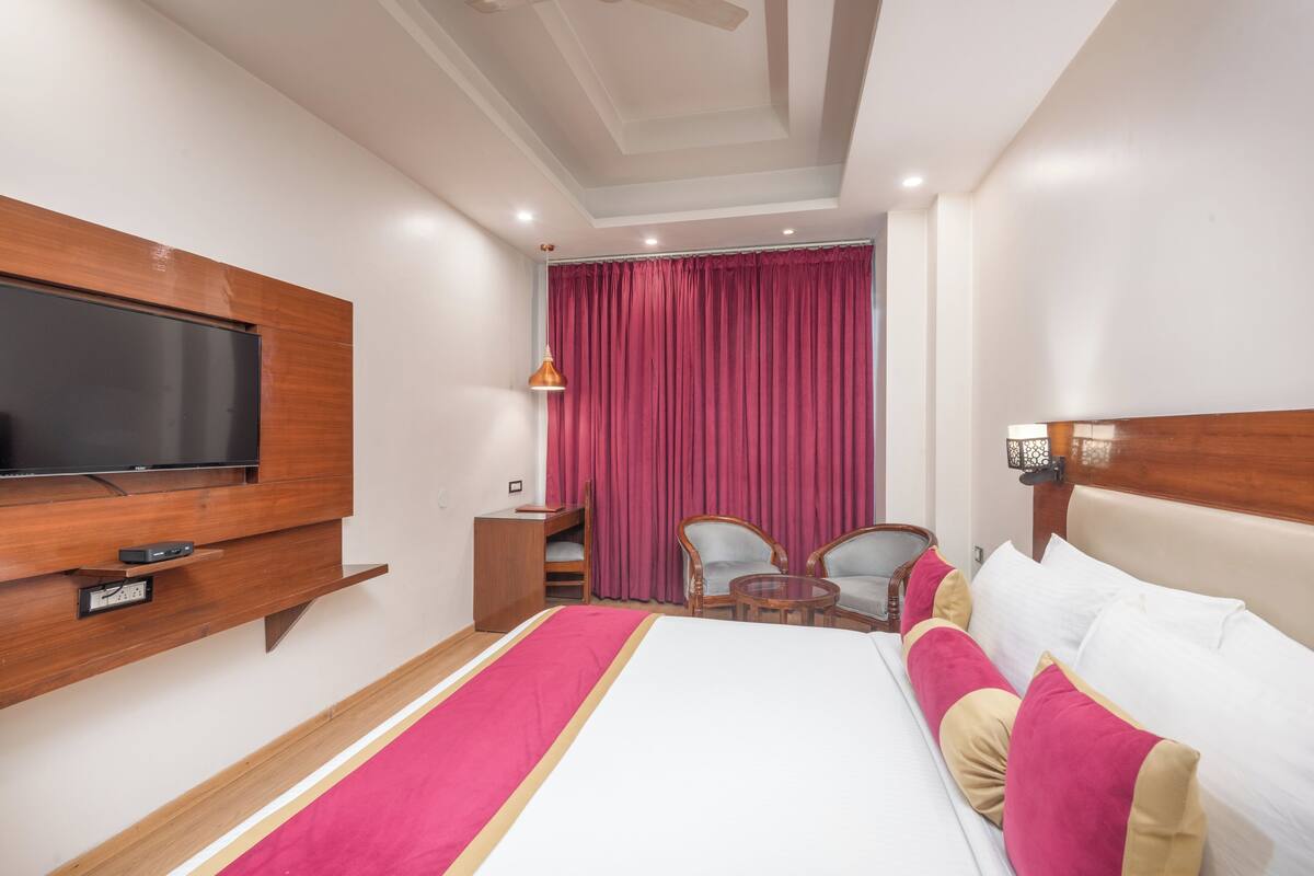 A hotel room with a large bed, a TV mounted on the wall, a desk with a lamp, two chairs, and a window with red curtains.