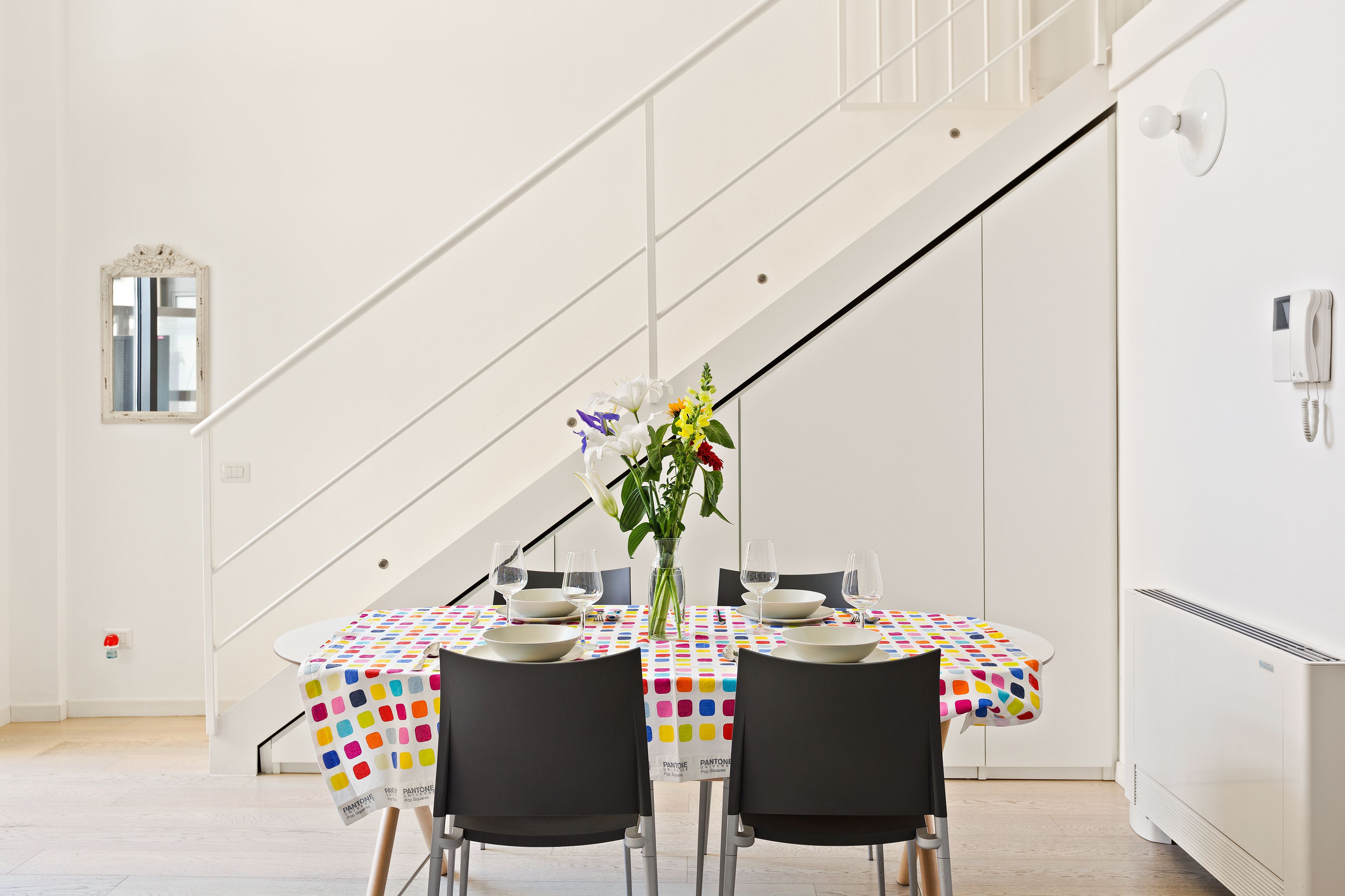 A dining area with a table set for two, a vase with flowers, and a staircase in the background.