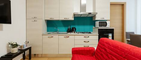 A compact kitchen with white cabinets, a teal backsplash, and a red couch in the living area.