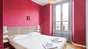 Un dormitorio con cama, ropa de cama blanca, una pared roja y un balcón con ventana.
