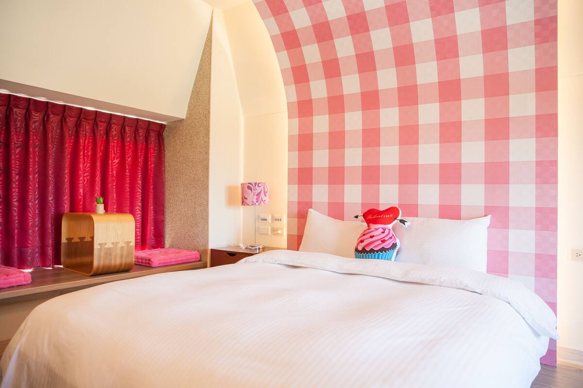 Habitación de hotel con una cama grande, una mesita de noche de madera, cortinas rojas y una pared con estampado rosa a cuadros.