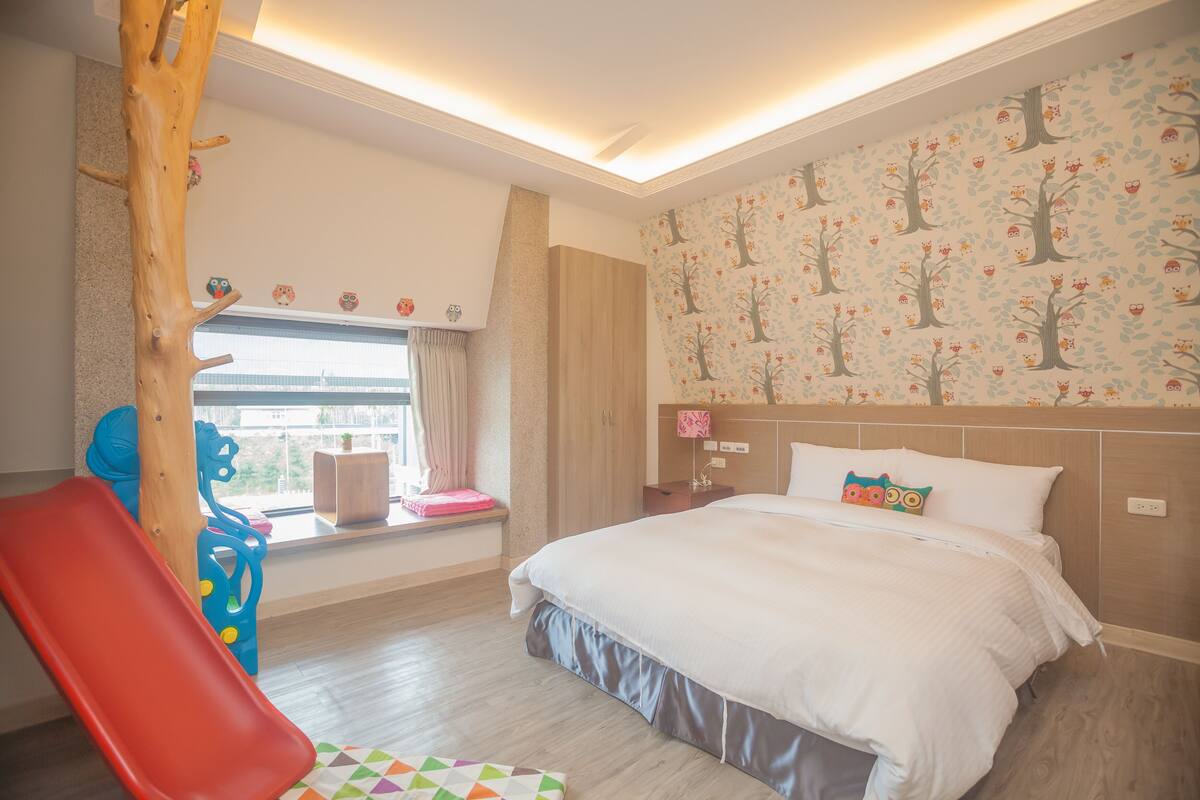 Un dormitorio con cama, ventana con cortinas, adorno de árbol de madera y zona de juegos colorida.