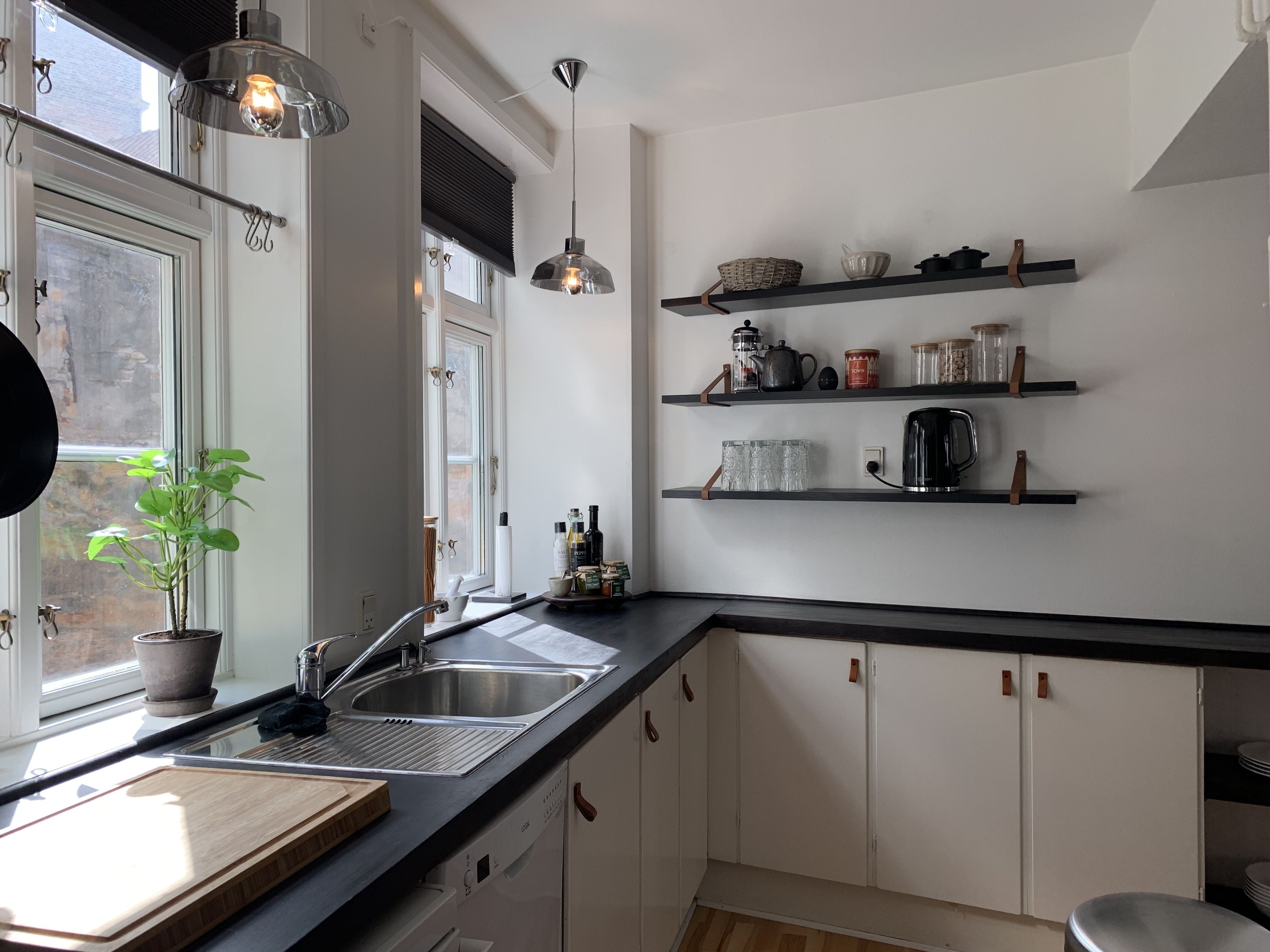A modern kitchen with a large window, stainless steel sink, and black countertops. There are shelves above the sink holding kitchen items, and a potted plant on the windowsill.