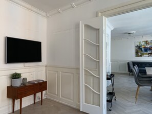 Un salon moderne doté d’un téléviseur à écran plat, d’une console en bois, d’un mur blanc agrémenté de moulures décoratives et d’une porte-fenêtre coulissante donnant sur une autre pièce.