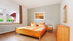 Une chambre à coucher avec un lit en bois, une table de chevet, une commode, une fenêtre avec des rideaux et un tableau encadré accroché au mur.