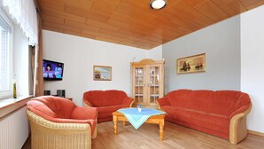 Un salon avec deux canapés rouges, un fauteuil en osier, une table basse en bois et un téléviseur fixé au mur.