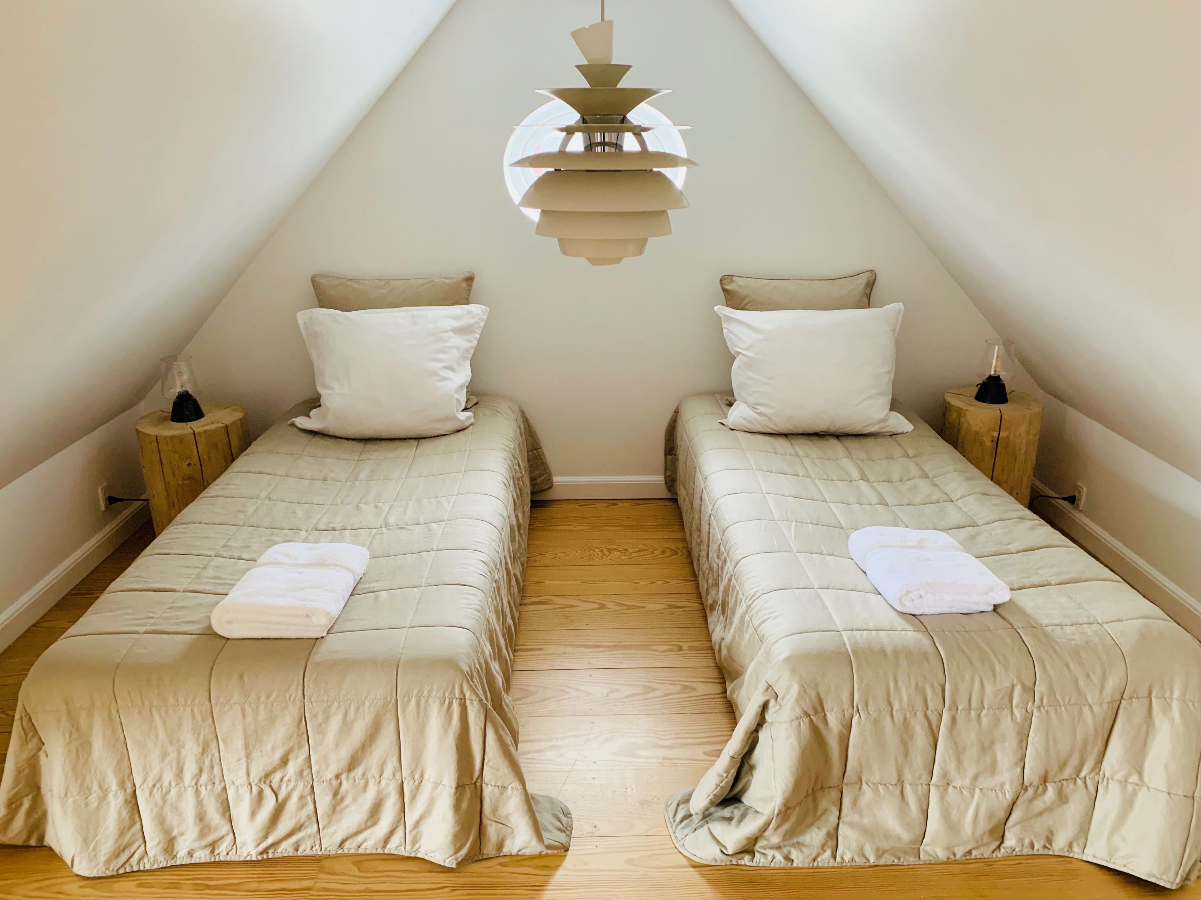 A room with two beds, each with a pillow and a white towel, wooden nightstands, and a modern pendant light.