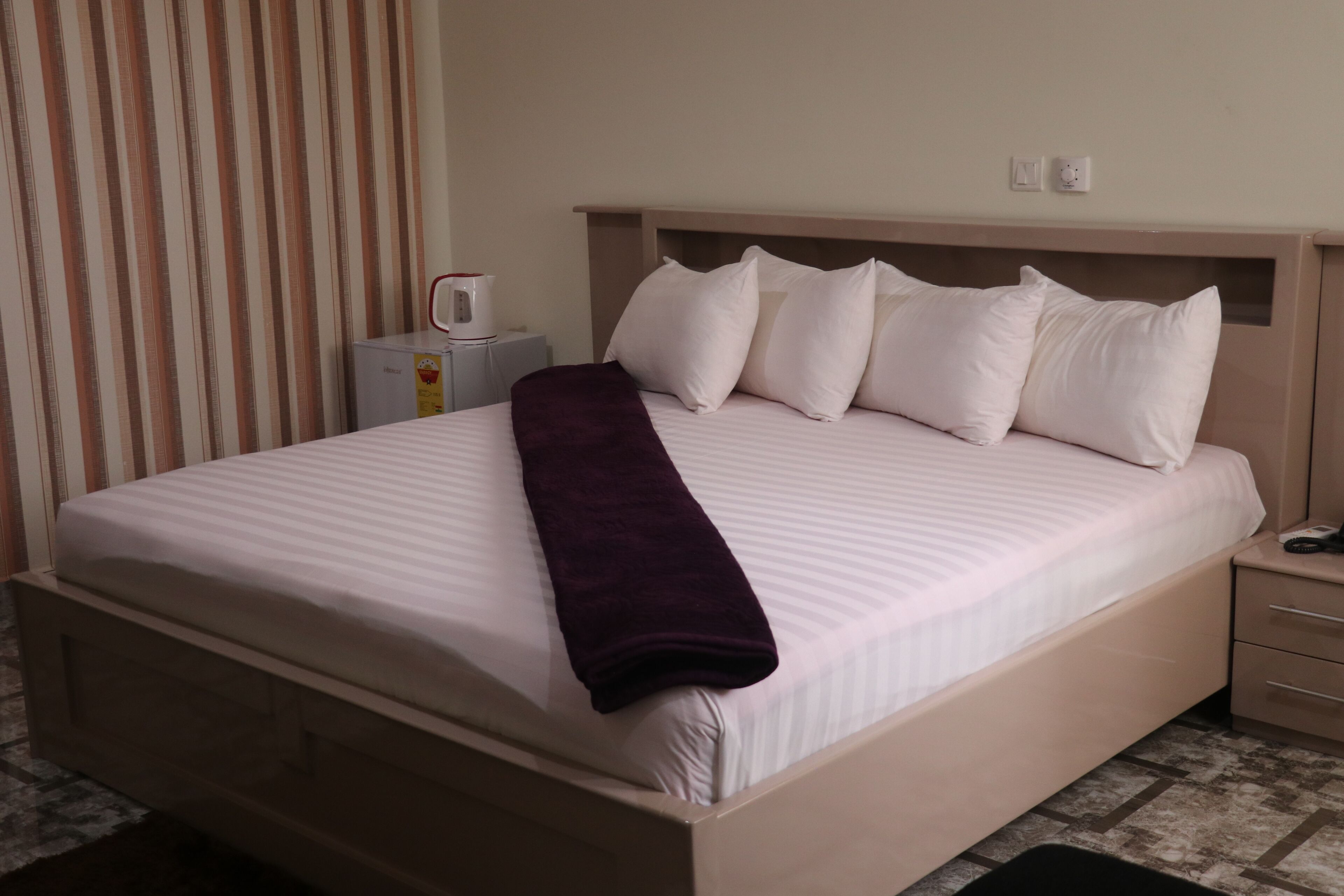 A neatly made bed with a white comforter and a purple throw blanket, a bedside table with a lamp, and a striped curtain in a hotel room.
