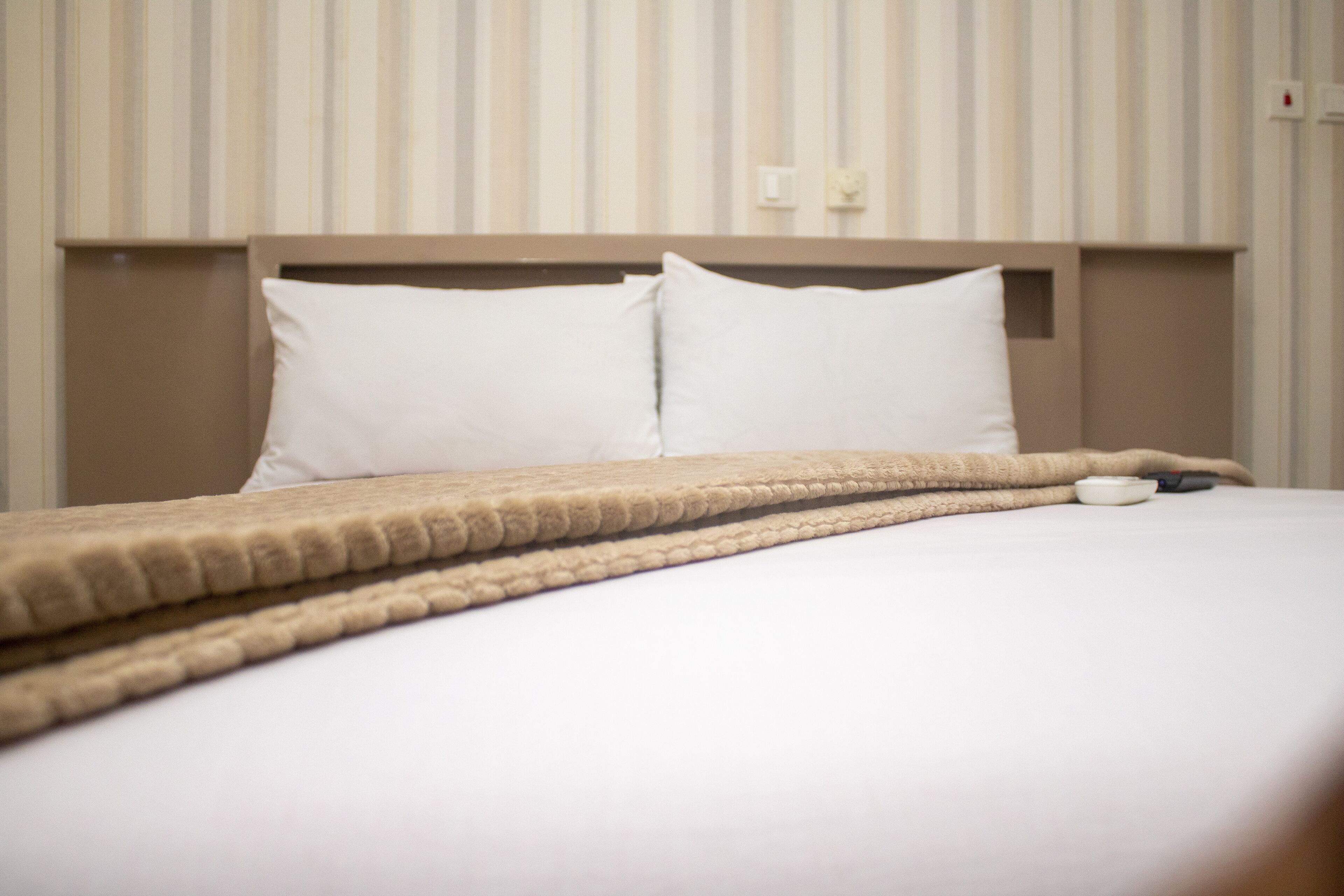 Una cama bien hecha con una manta color beige y dos almohadas, una mesita de noche con un control remoto y una pared con rayas verticales.
