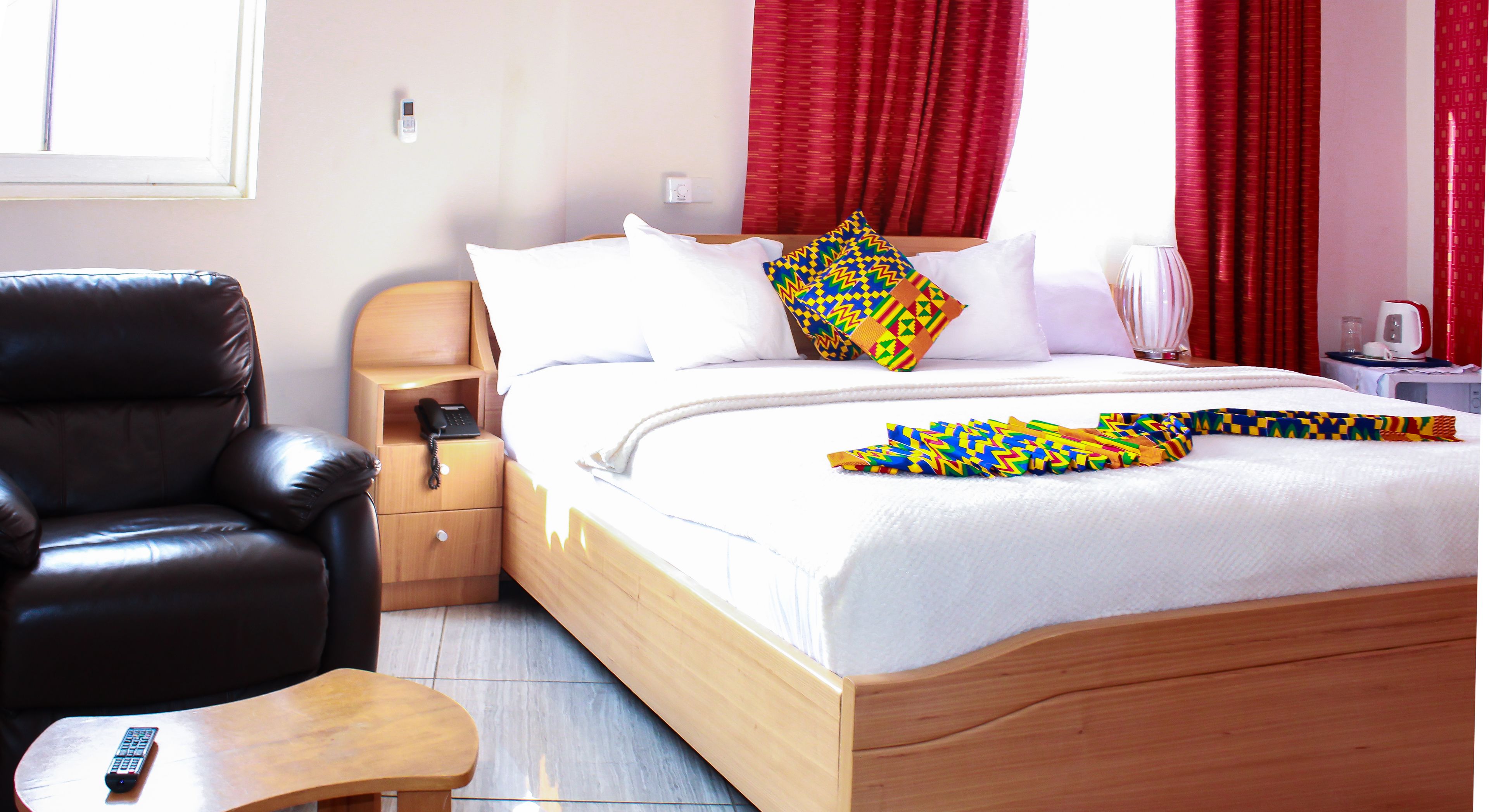 A hotel room with a wooden bed, a leather chair, a bedside table, and a red curtain.