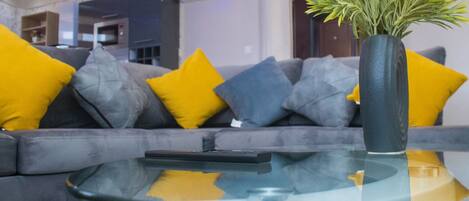 Una sala de estar moderna con un sofá gris, cojines amarillos y grises, y una mesa de centro de vidrio con un control remoto.