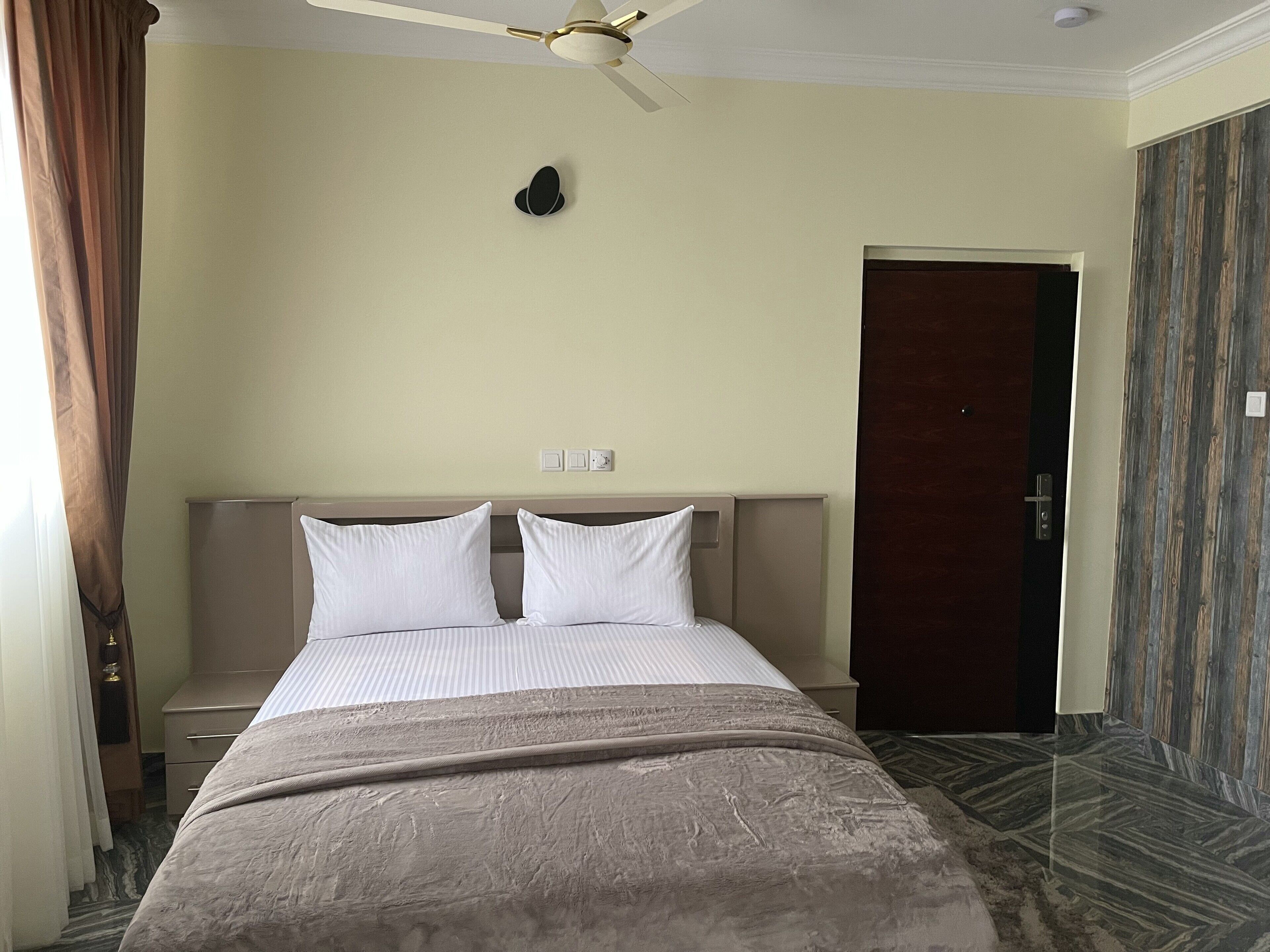 Ein Hotelzimmer mit einem Bett, einem Nachttisch, einem Deckenventilator, einer Tür und einer Wand mit ornamentalem Wandpapier.