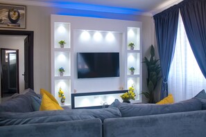 A modern living room with a large flat-screen TV mounted on a wall, grey sofas, yellow and grey cushions, potted plants, and a large window with curtains. - Exotic Palace Hotel (Accra)