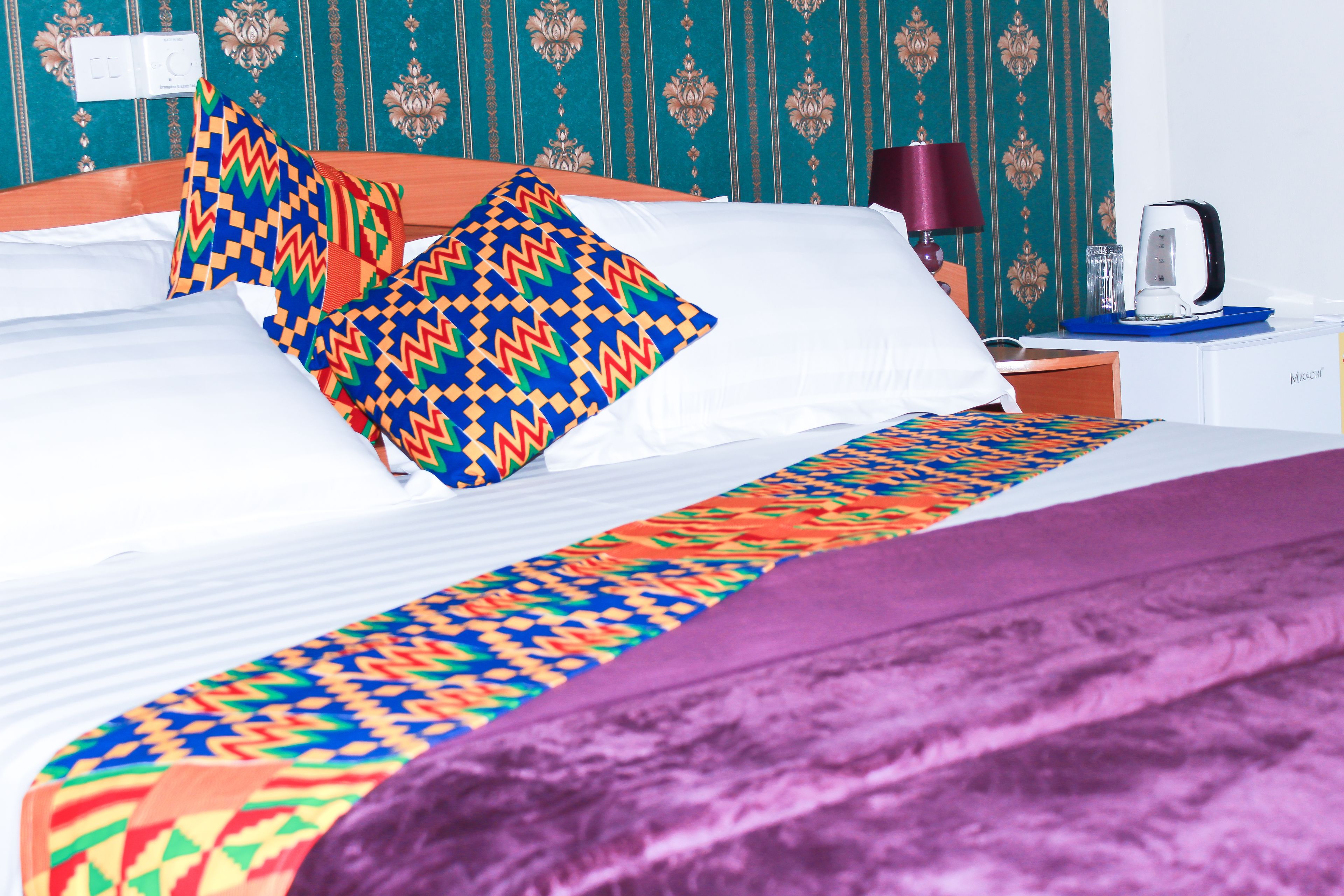 A neatly made bed with colorful patterned pillows, a purple throw blanket, and a nightstand with a lamp and a coffee maker.