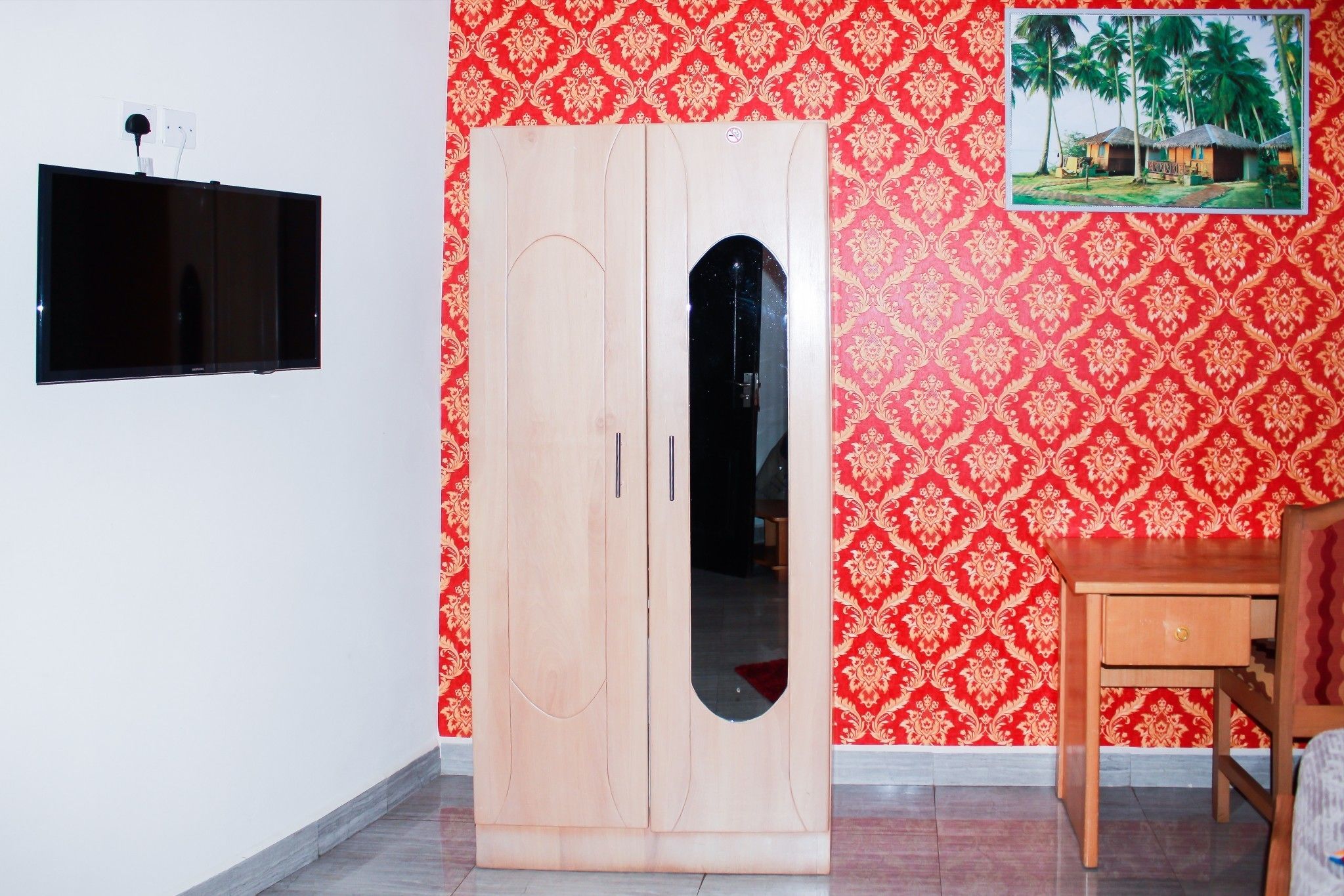 A room with a wooden wardrobe, a television mounted on the wall, and a patterned wallpaper.