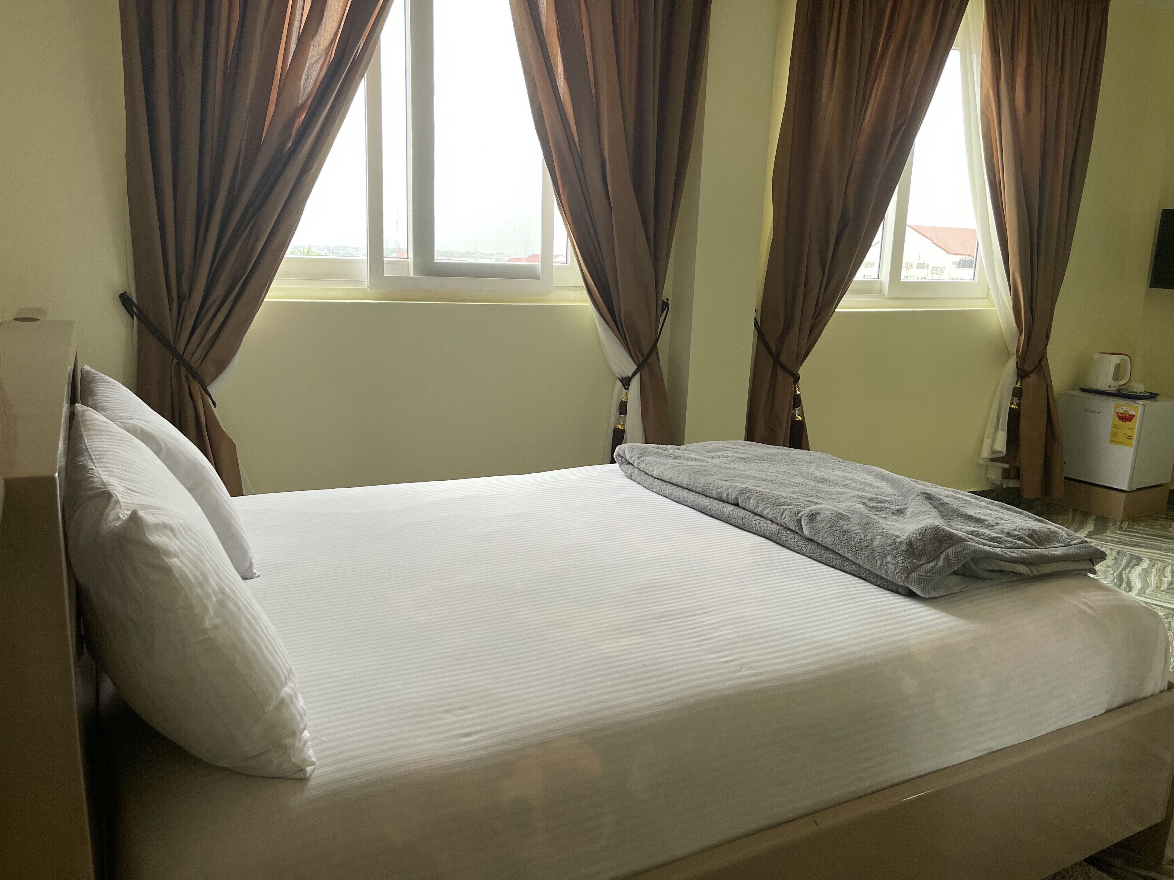 A neatly made bed with white linens and a gray blanket, curtains partially drawn, and a small refrigerator with a coffee cup on top.