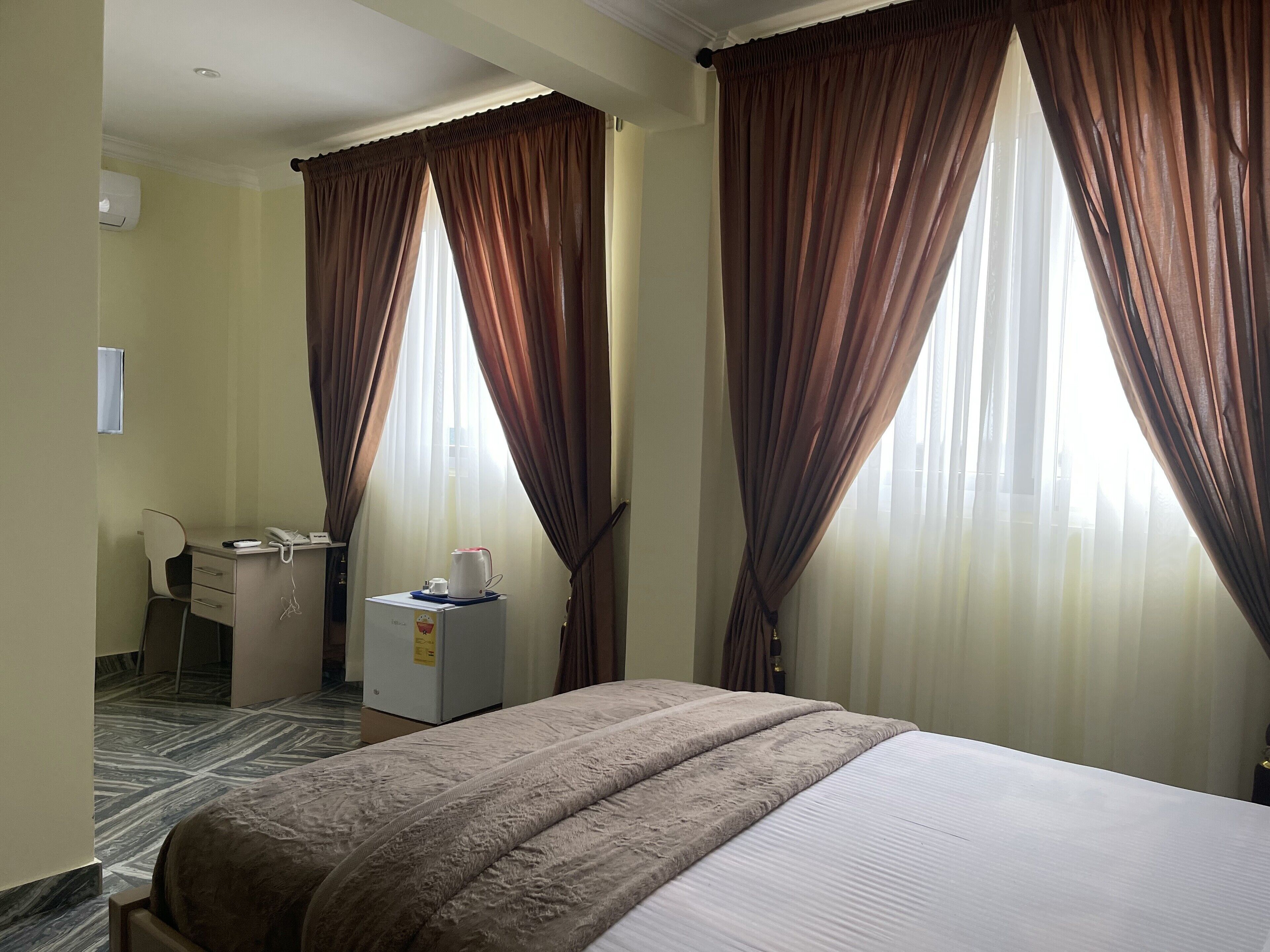 A hotel room with a bed, a desk with a chair, a small refrigerator, and curtains.