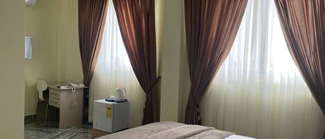 A hotel room with a bed, a desk with a chair, a small refrigerator, and curtains.