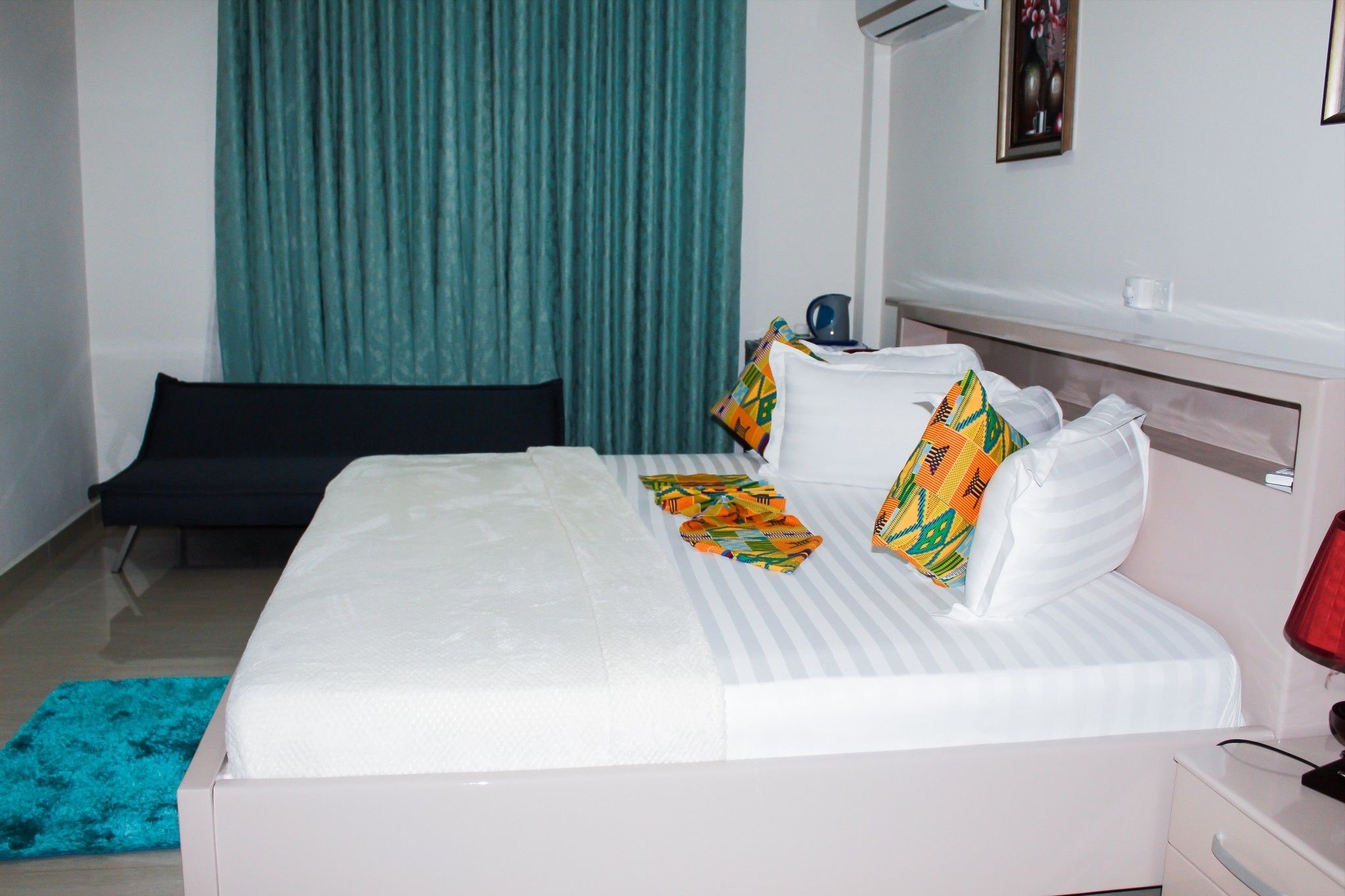 A neatly made bed with white linens and colorful pillows, a black bench, a teal curtain, and a small table with a red lamp.