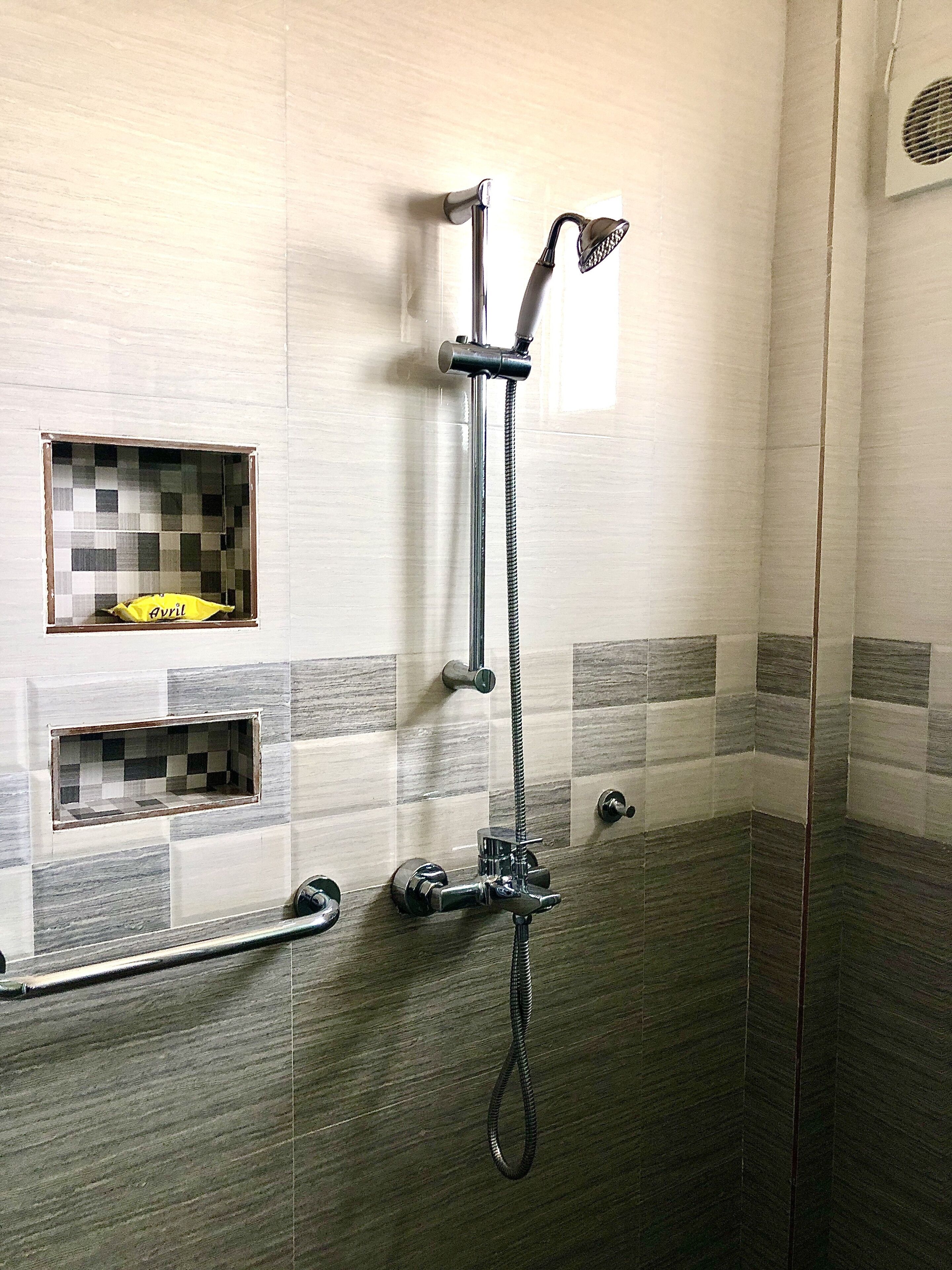A shower with a handheld showerhead and a fixed showerhead, a grab bar, and a small window with a yellow bag labeled "Avril".