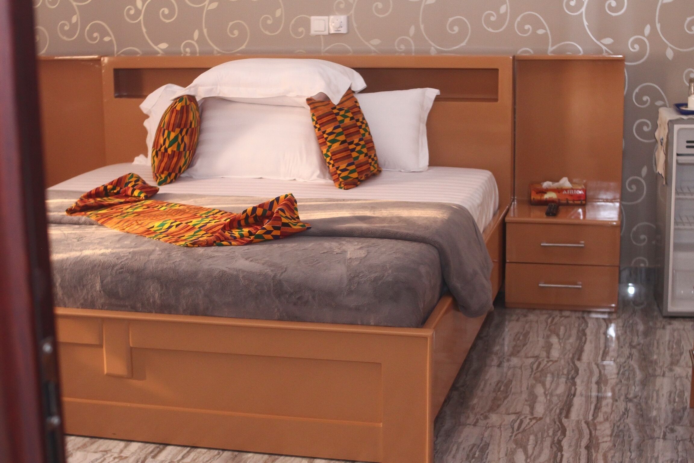 A neatly made bed with patterned pillows and a blanket, a wooden nightstand with a remote control, and a small cabinet with a tissue box.