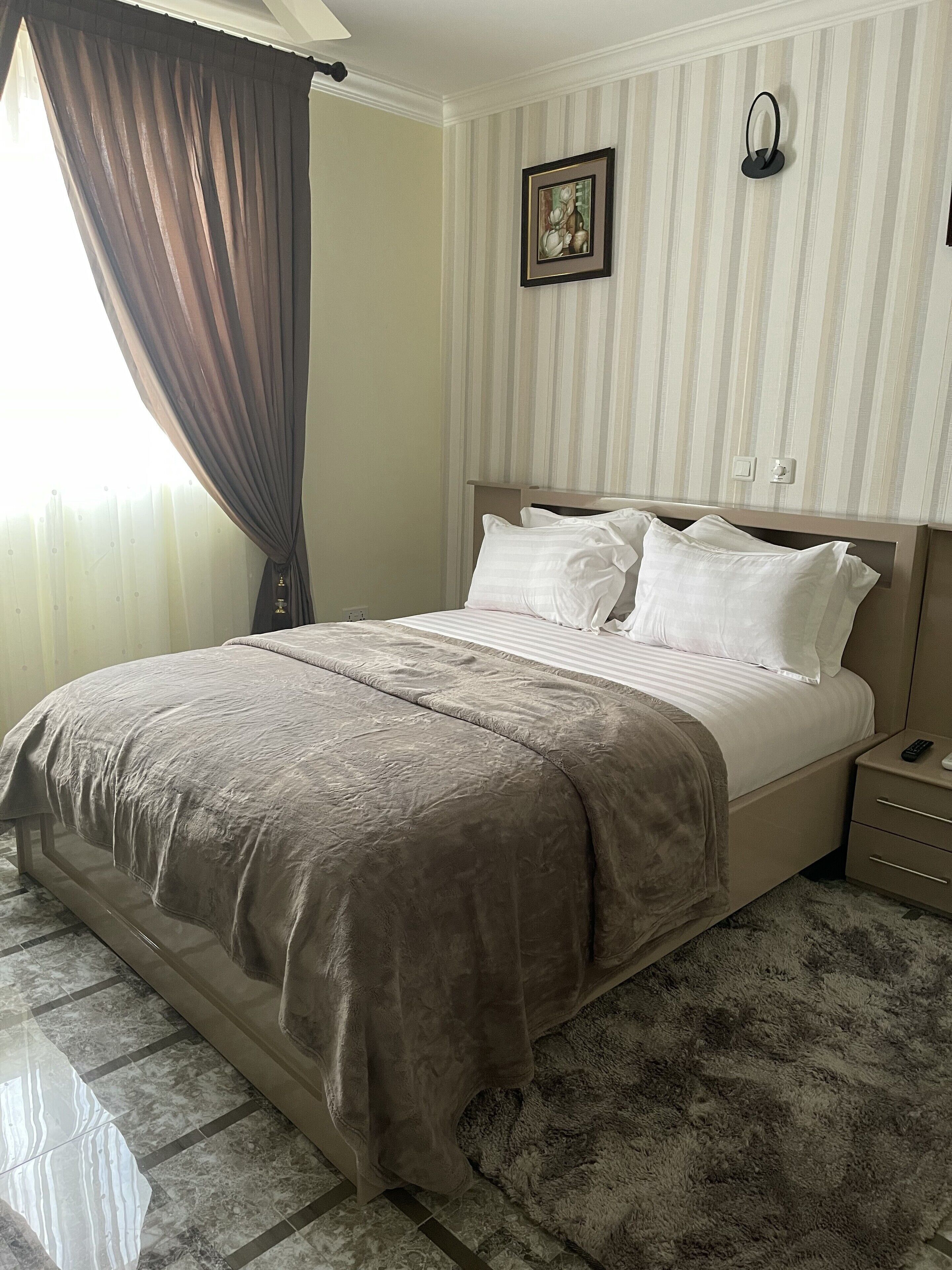 Una cama bien hecha con una colcha marrón y almohadas blancas, una mesita de noche con un control remoto, y un cuadro en la pared.