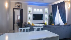 A modern living room with a grey sofa, white dining table, and a flat-screen TV mounted on the wall. - Exotic Palace Hotel (Accra)